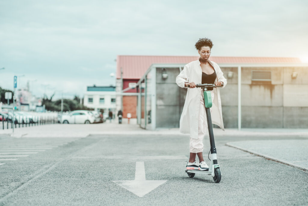 A young African fancy female in glasses and a white cloak is riding an e-scooter with green accumulator on the asphalt road with a painted arrow as a road marking, sunny day, shallow depth of field