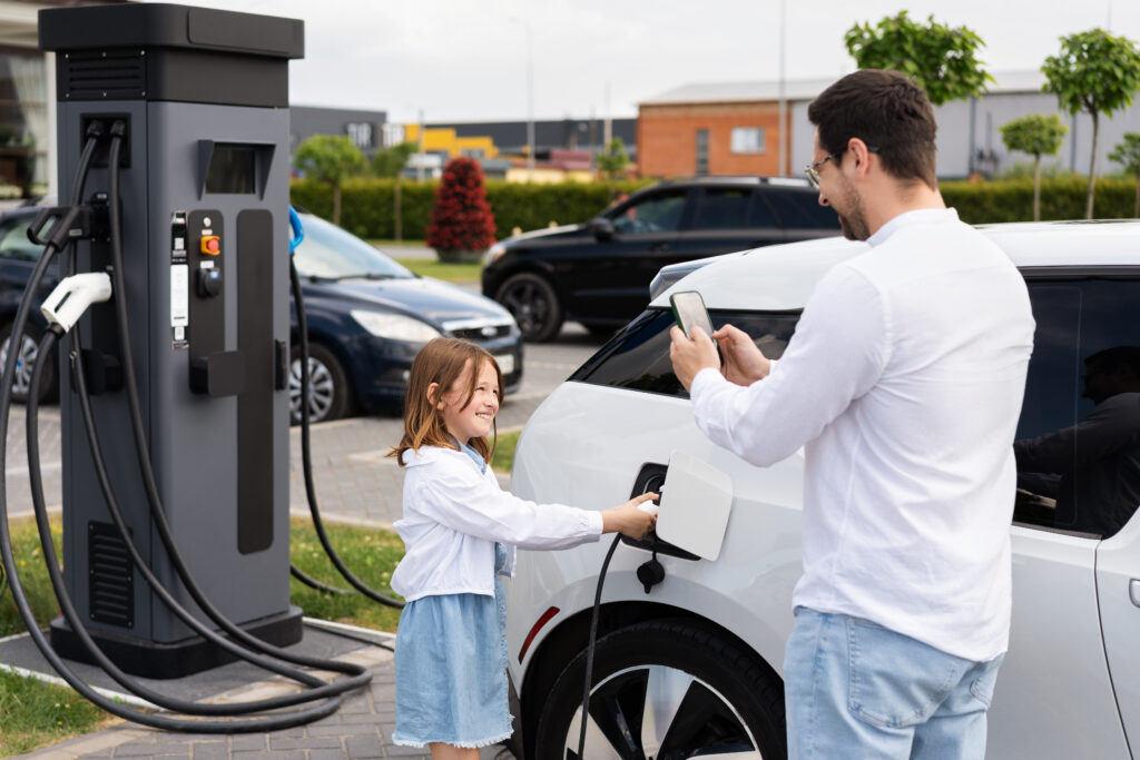 Father And Daughter Charging Electric Car At Station Using Smartphone